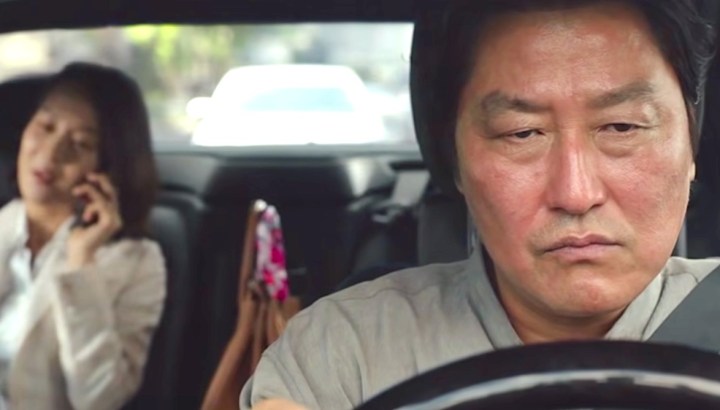 Song Kang-ho grimaces behind a steering wheel as Cho Yeo-jeong talks obliviously on the phone in the backseat in a still from the movie Parasite.