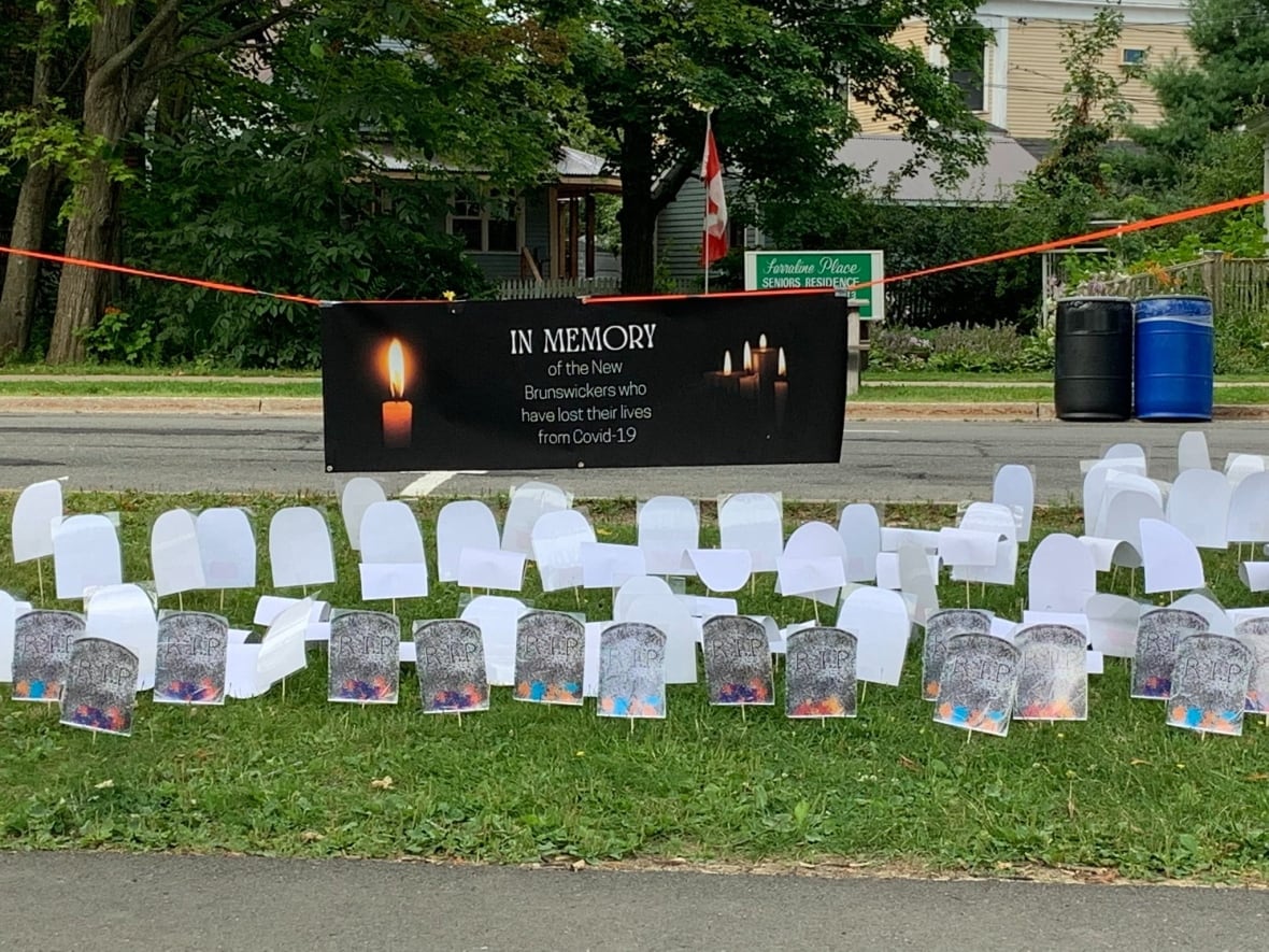 Paper memorial tombstones are plucked into the grass in front of a road. A black banner hangs above the tombstone. It shows lit candles, and reads 'in memory of the New Brunswickers who have lost their lives from COVID-19.'